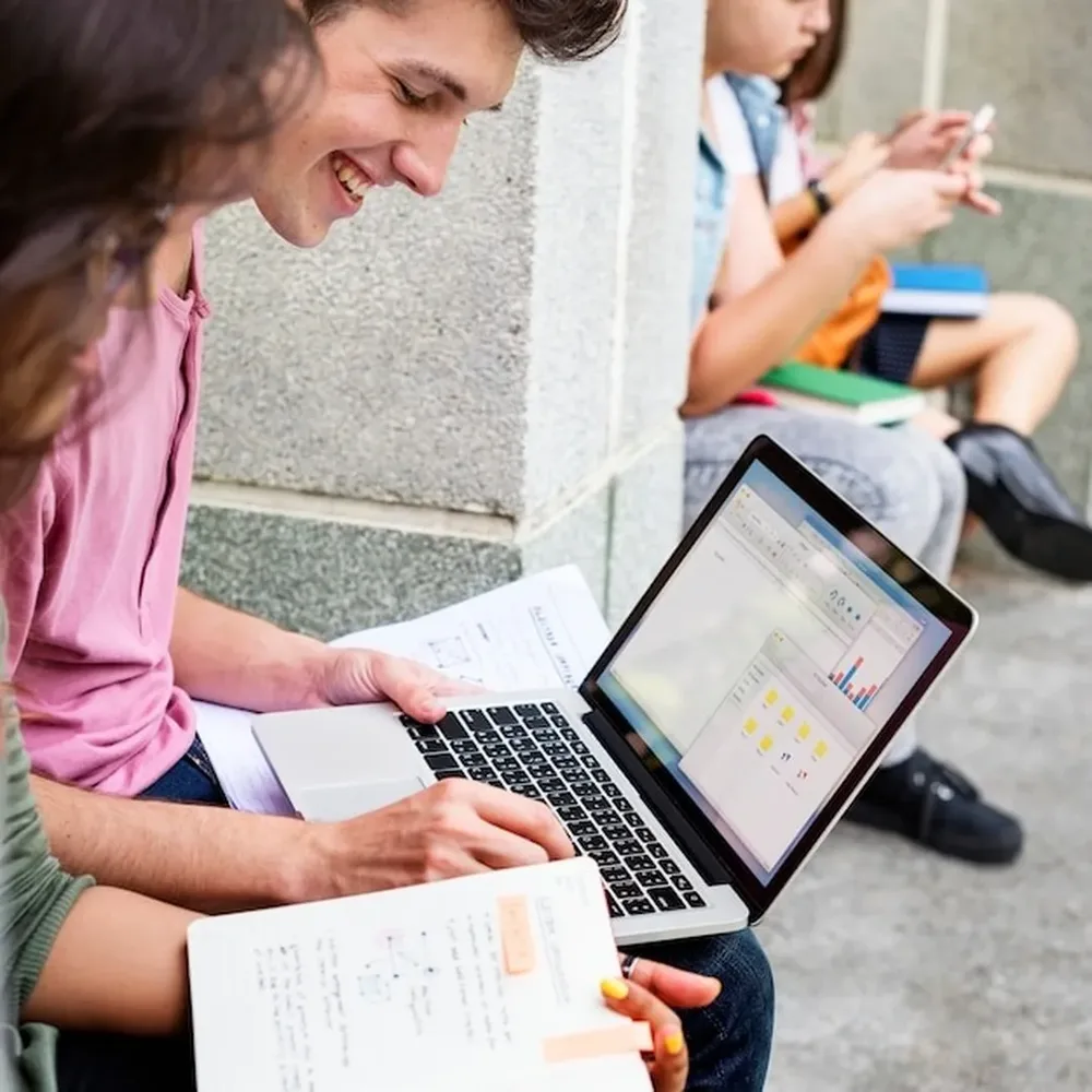 A student working on a laptop with notes while classmates study nearby