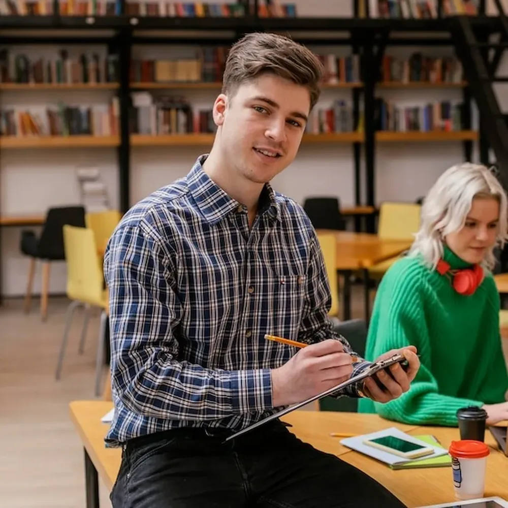 A student taking notes in a university library with a classmate behind him