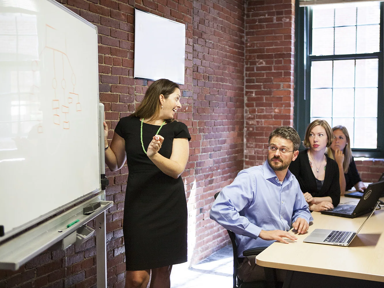 A project lead walks a localization team through a workflow diagram on a whiteboard while teammates follow along on their laptops